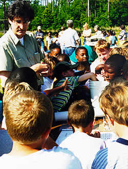 Photo: young students crowd around  as an adult  speaks to them. at an outdoor science fair.