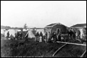 Athapascan women & children pose in front of cabins and tents with miners, Circle, Alaska, 1895