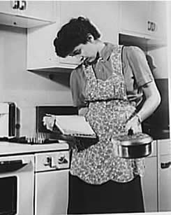Woman holding and reading a cookbook, and holding a saucepan.  The burner on the gas stove is lit.