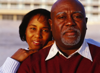 A man and a woman standing.  The middle-aged man is in the foreground, looking into camera.  The woman is standing behind him with her hands on his shoulder, smiling and also looking into the camera. 