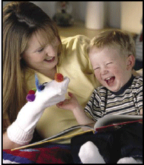 Photo of a woman reading with a laughing child; the woman is wearing a sock puppet on her hand