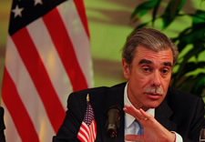 Secretary Gutierrez gesturing in front of microphone with American flag in the background.