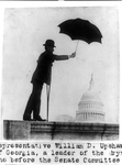 [Representative William D. Upshaw of Georgia, a leader of the drys, standing on railing with crutch and holding umbrella, with dome of the U.S. Capitol under the umbrella
