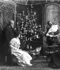 Photo:  three people. one holding a baby, near a Christmas tree with lighted candles on it.