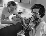 Alan Jabbour playing the fiddle in a kitchen area, with Dean Reed holding a phone up to the fiddle