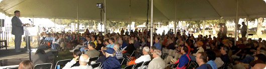 A man speaking at a podium, to a large crowd of people under a tent.