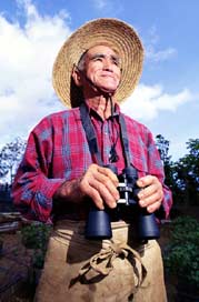 Smiling man standing outdoors, wearing a sunhat and holding binoculars