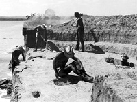 Image of Archeologists excavating near rising resevoir waters.