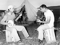 Image of  two men, one playing the banjo and the other playing the guitar in a feild camp.