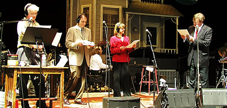 From left to right,  Fred Newman, Tim Russell, Sue Scott, and Garrison Keillor perform a skit from (ital) A Prairie Home Companion (close ital) against a backdrop featuring a housefront and vintage advertising signs. Each perfomer stands in front of  a stand microphone, and to the far left there is a sound effects table.