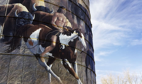 Detail of a mural painted on a water tank.  A barechested Native American brave, rides a brown and white horse, carrying a bow in his right hand. A herd of buffalo follows behind