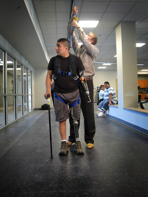 This photo shows a wounded Army National Guard member who had his left leg and arm amputated, practicing walking in a corridor.  He is being hooked into a harness to prevent him from falling.  