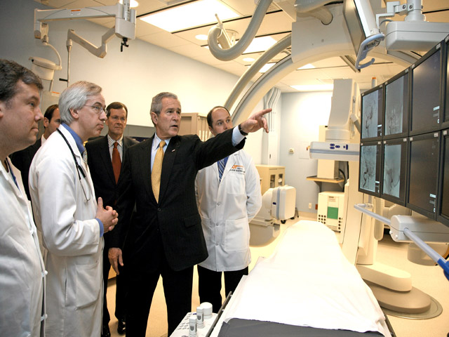 A photograph of President Bush touring a hospital with some doctors, pointing at digital radiology equipment.