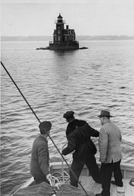 Photo of three men in a boar in foreground, with lighthouse , surrounded by water, in distance.