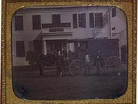 View of a horse and covered cart in front of white frame building with signs for County House, J. Griggs and L.J. Phillips, Daguerrian Rooms, Thompson, Connecticut
