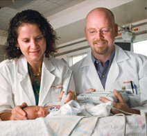 The photograph shows two doctors monitoring a newborn in a neonatal intensive care unit.