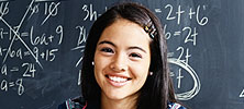 A girl, smiling, sitting in front of a chalkboard.
