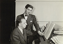 Bernstein reads music over the shoulder of Copland, seated at piano