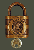 An historical picture of an old Forest Service lock and key.  The words "Forest Service" and "US" are engraved on the lock, with a engraved pine tree separating the "U" and "S".  A small key is also inserted in the bottom of the lock.