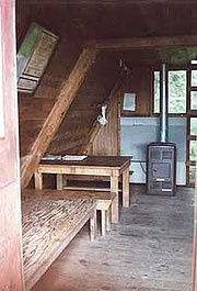 The inside of a typical A-frame cabin showing a wooden platform bunk, wood table and bench, and an oil-fired stove under the sloping cabin side, with the ladder to the loft visible near the stove.