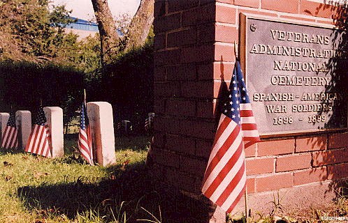 Photo of a iron plaque mounted on a small brick wall stating that veterans of the Spanish American War resides in that cemetery. Several upright markers with small American flags are mounted in front of each headstone.