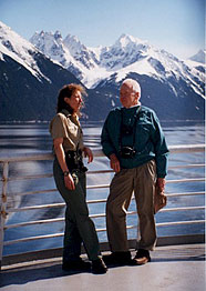 Forest Service intepreter on the outside deck of an Alaskan Ferry, talking to a passenger with snowcapped peaks in the background.