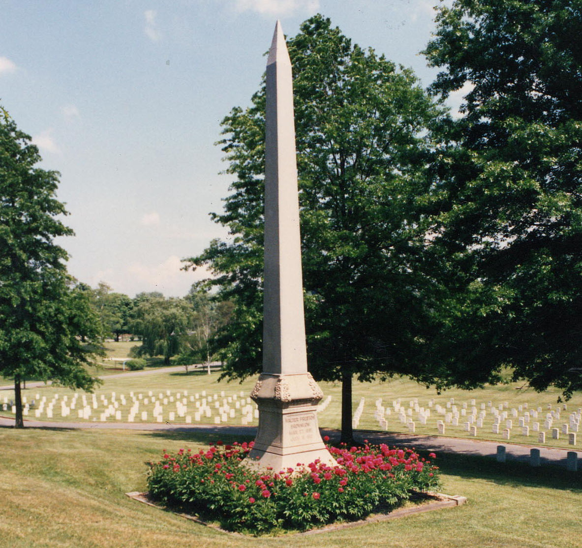 A photo of a stone needle point monument with flowers placed at the base. Upright markers are viewed in the far distance.