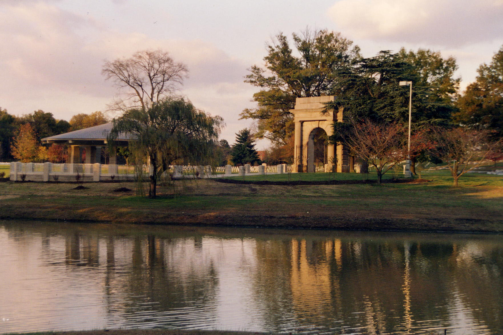 A photo of a lake reflecting a committal shelter located behind a small willow tree next to a stone archway with upright headstones in the distance.