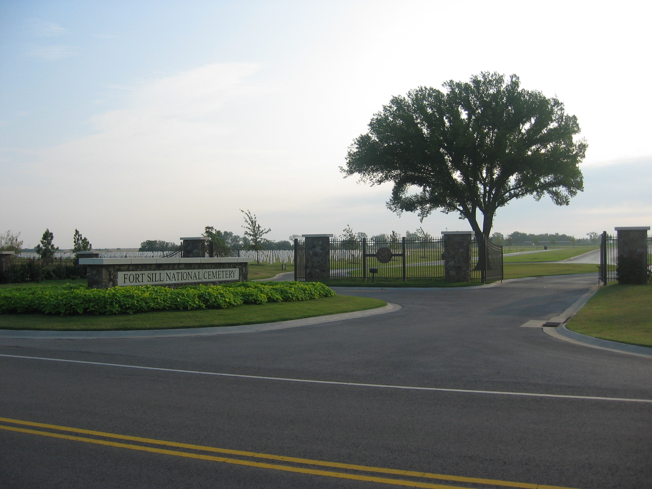 A photo of Fort Sill's entrance gate. A rectangular cobble-stone wall that stands alone on an oval-shaped grass meridian.  Opened iron gates leads into the cemetery with upright markers shown in the background.