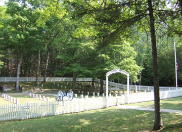 Overall view of Fort Mackinac Post Cemetery showing the entrance gate and white picket fence that surrounds the lot. Within the fence are headstones, each marked with a miniature flag, and a cannon on a wooden carriage.