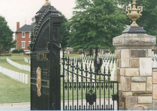 A photo of an iron gate with upright markers in the background along with a partial view of a colonial brick house in the background.