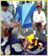 Photo of a family gathered around a campfire roasting marshmallows. Their tent is in the background.