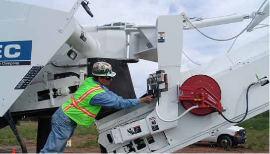 A Wirtgen pavement-milling machine removes the top layer of asphalt from a former airport runway in Marquette, Mich., during controlled field testing of prototype water-spray dust-suppression systems in June 2008, as NIOSH investigator Li-Ming Lo, of DART, walks alongside.  NIOSH wind-measuring equipment and video equipment can be seen mounted on the top of the machine.