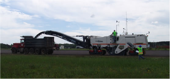A Wirtgen pavement-milling machine removes the top layer of asphalt from a former airport runway in Marquette, Mich., during controlled field testing of prototype water-spray dust-suppression systems in June 2008, as NIOSH investigator Li-Ming Lo, of DART, walks alongside.  NIOSH wind-measuring equipment and video equipment can be seen mounted on the top of the machine.