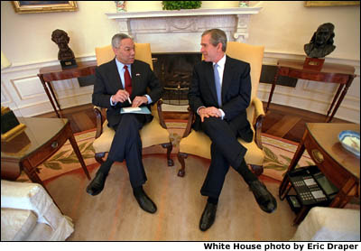 President Bush and Secretary Colin Powell sit in yellow chairs in front of the Oval Office fireplace. White House photo by Eric Draper.