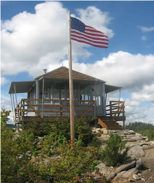 photo of Gold Butte Lookout from trail leading up to it. American flag in front. Photo shows hip roof construction with catwalk surrounding and glassed walls