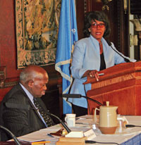 A woman speaking at a podium while a man listens.