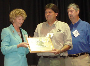 Photograph: Gail Tunberg presenting award to Gary Rotta; Rocky Evans looks on. Award is an ingraved plaque.