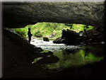 A hiker stands upright on the bank of the underground stream flowing out of the mouth of the Cavern Lake cave.