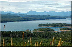 A panoramic view from a hillside shows forested lowlands, open waterways, islands, and mountains in the background.