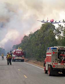 photo: Air tanker flying through smoke; fire trucks and firefighters on road below.