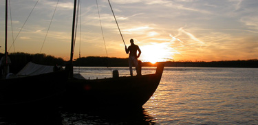 Man on keelboat with setting sun behind.