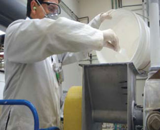 A nanoparticle production worker wearing a personal air sampler and hearing, respiratory, and dermal protection during a pouring operation.