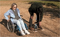 Picture of a woman in a wheelchair and a man standing beside her on a playground surface covered with wood chips.