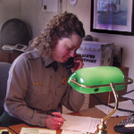Park employee sits at desk under green lamp, working on open mail and paperwork while talking on the phone.
