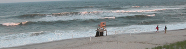 Tinted by the glow of the setting sun, couple strolls along beach behind unoccupied lifeguard stand, as frothy waves break on the shore.