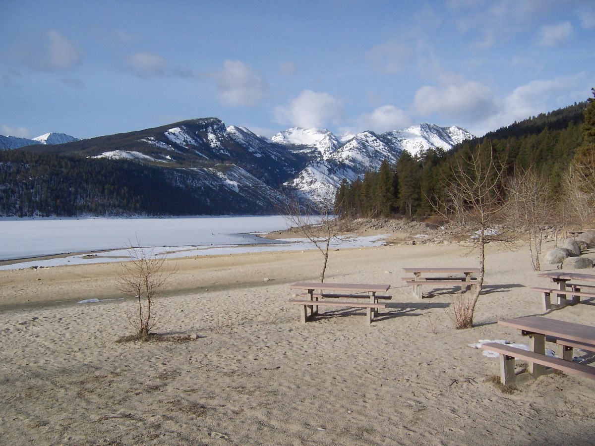 Awaiting summer at Lake Como beach.  Snowcapped mountains in the background.  Sandy beach area in the foreground with picnic tables and small tree's.  Photo courtesy of  Les Myers, Recreation Technician - Darby Ranger District.