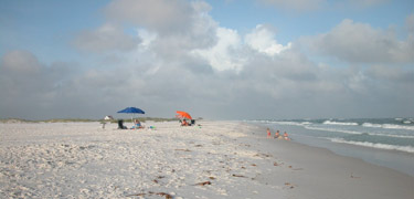 This is a beach scene with a white, sandy beach, visitors under brightly colored beach umbrellas, rolling surf, and puffy clouds.