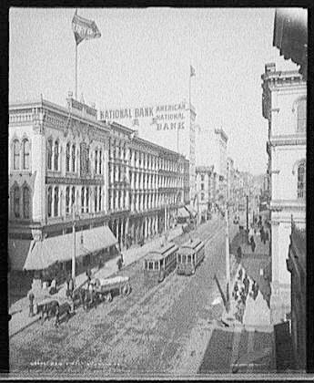 Larga vista de los edificios a lo largo de Main St. en Richmond Virginia, con dos carros de tranvía, un carruaje tirado por caballos y el American National Bank visible en la distancia.
