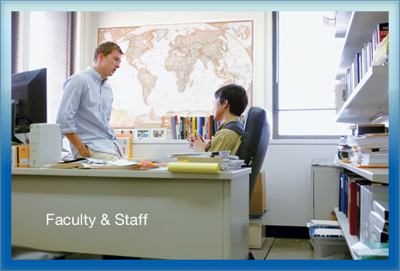 A faculty member seated at a desk talking to a  student standing in front of a world map